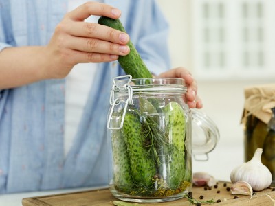 Woman putting cucumber into pickling jar at table in kitchen, cl