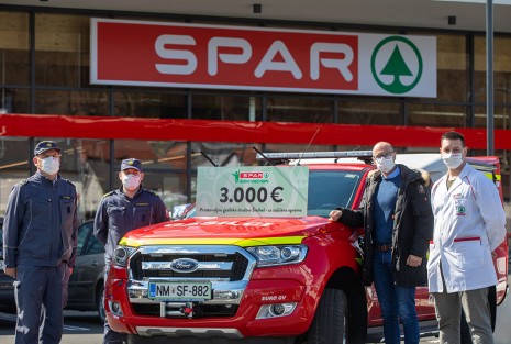 Dva gasilca ter dva predstavnika SPARA zraven gsailskega vozila na katerem je tabla z zneskom donacije 3000€ pred trvgovino SPAR