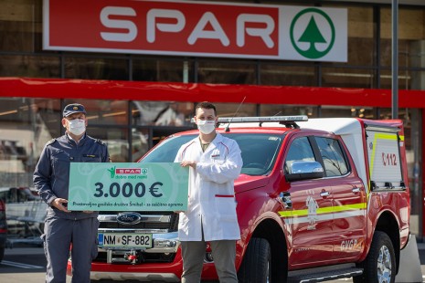 Gasilec ter predstavnik SPARA držida tablo z donacijo 3000€ pred gasilskim vozilom ter SPAR trgovino